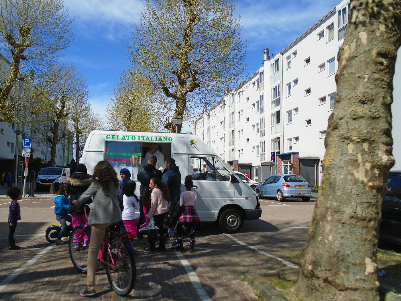 A van lettered Gelato Italiano parked on a street, children on bicycles passing it