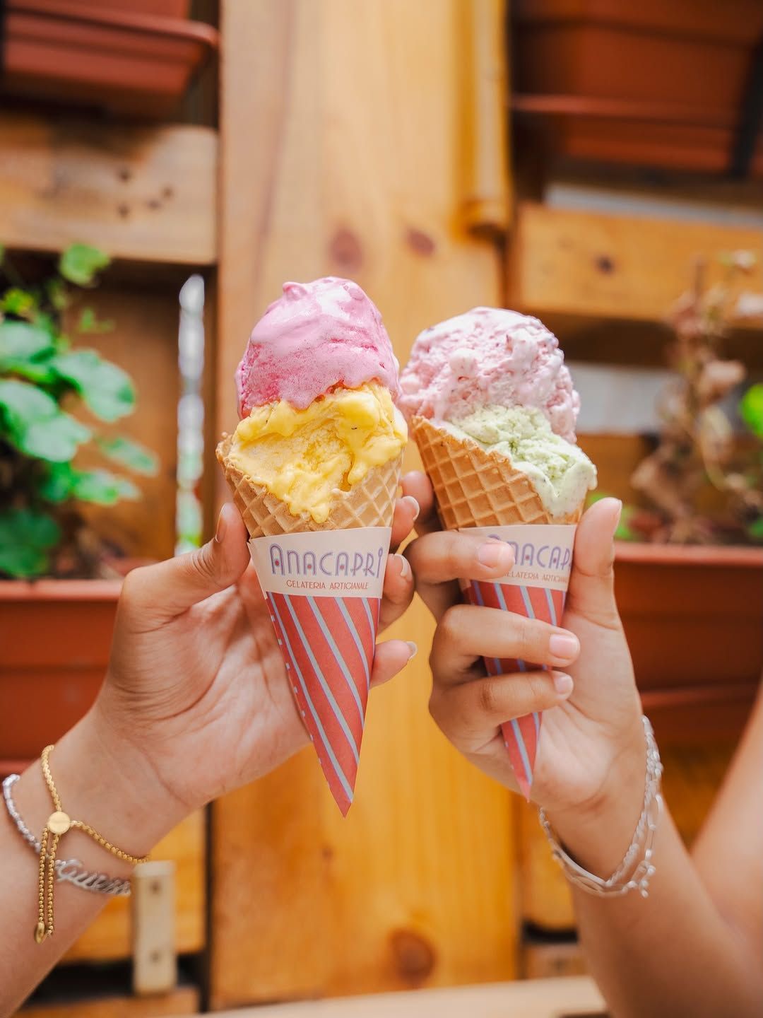 Two hands holding cones side by side, one pink over yellow, one pink over pistachio, in striped wrappers printed Anacapri Gelateria Artigianale