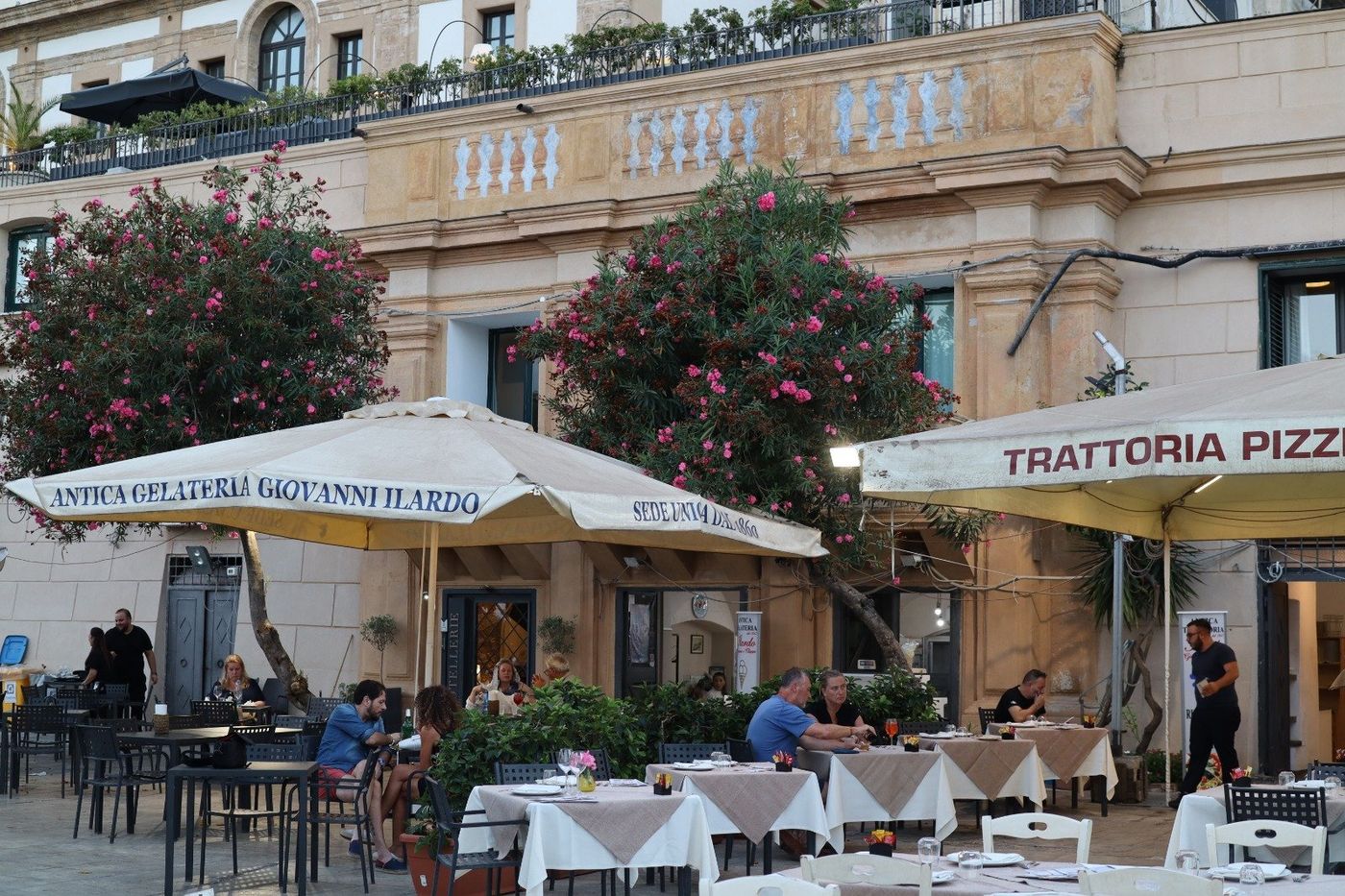A parasol lettered Antica Gelateria Giovanni Ilardo, sede unica dal 1860, over outdoor tables in front of a tall stone building, with oleanders in flower behind