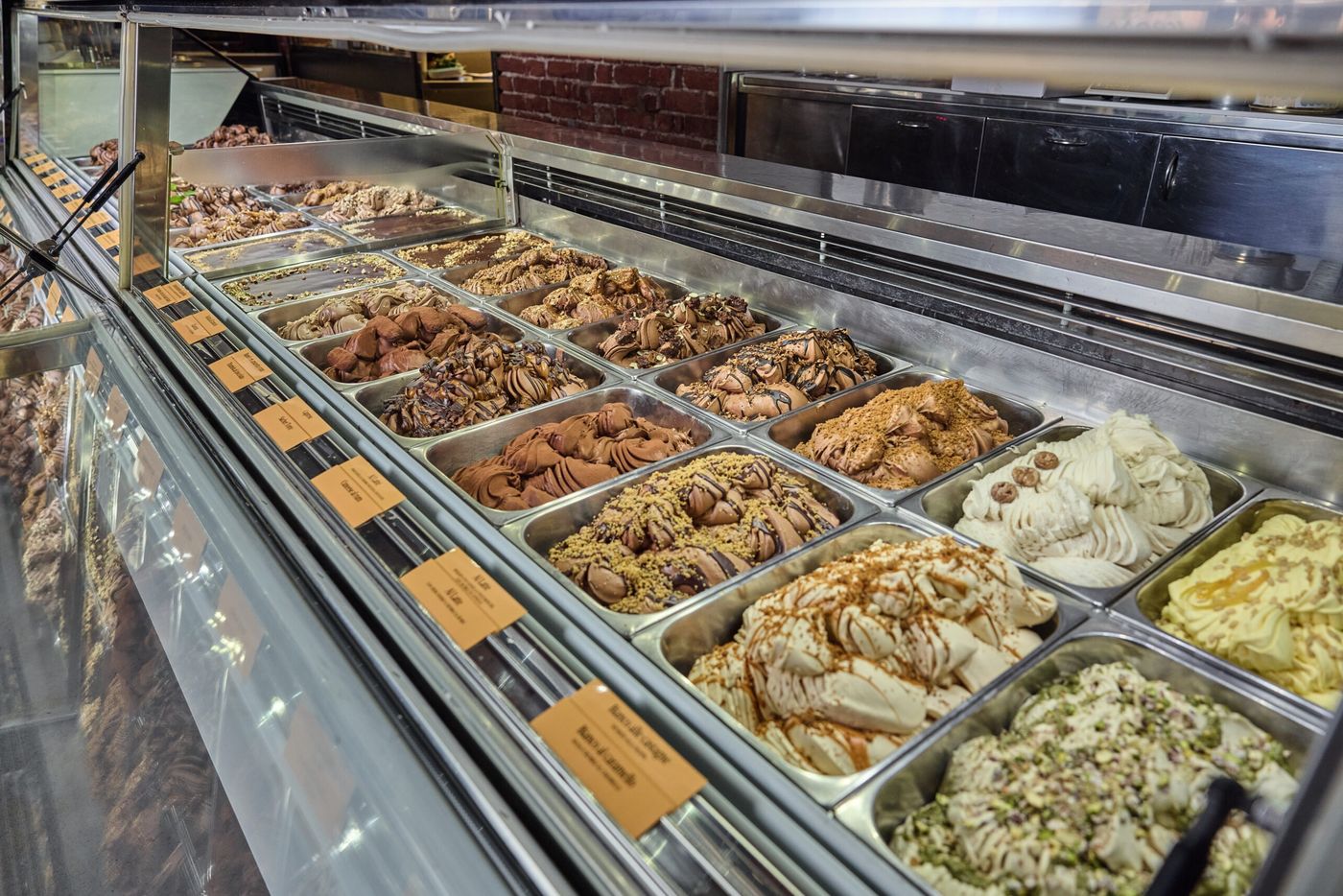 Metal trays of gelato in a lit display case, chocolate and pistachio and caramel flavours under small printed labels, with a brick wall behind the counter
