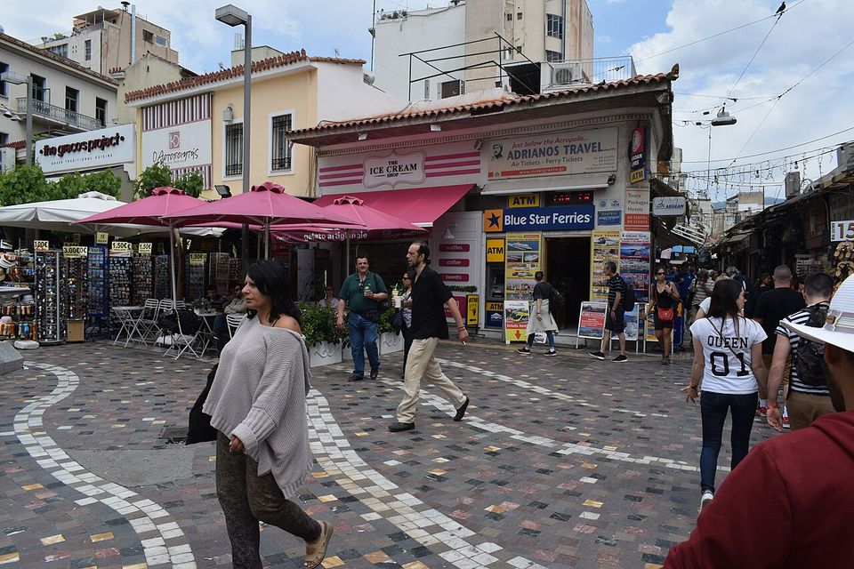 A gelato shop signed THE ICE CREAM SHOP under pink parasols on a busy paved square, people crossing in front of it