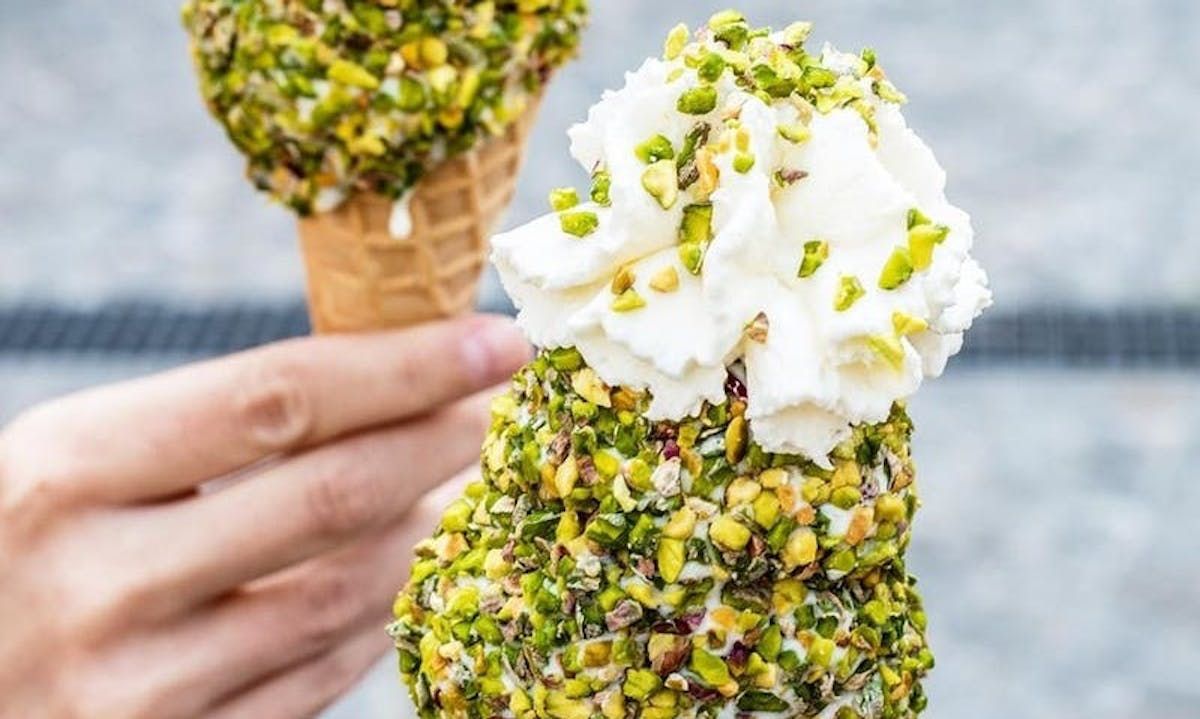 A hand holding two cones of pale ice cream rolled thickly in chopped pistachio