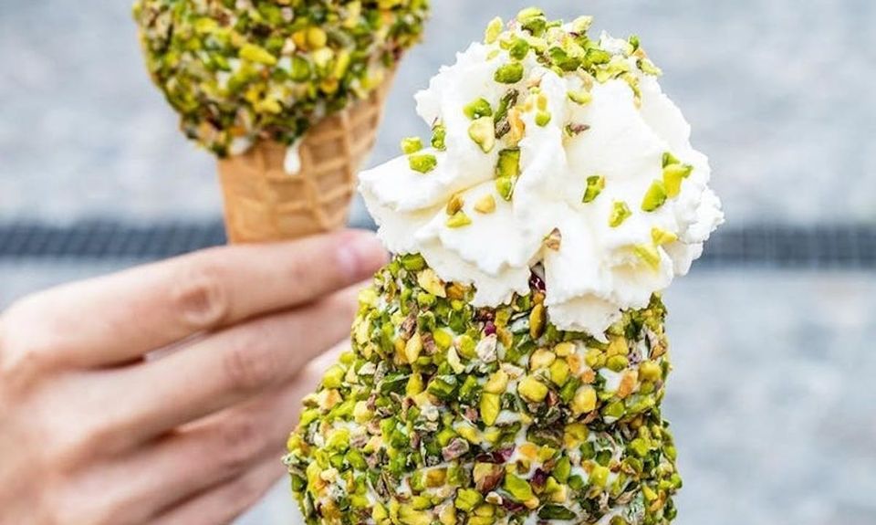 A hand holding two cones of pale ice cream rolled thickly in chopped pistachio