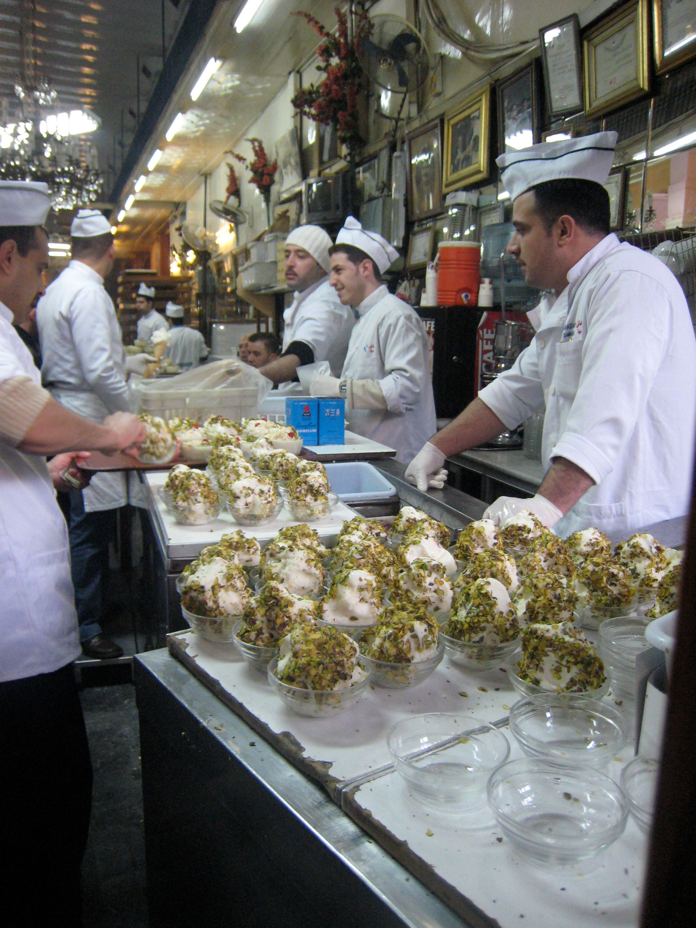 Staff in white coats and paper caps serving booza at the counter, bowls of it heaped with crushed pistachio along the front