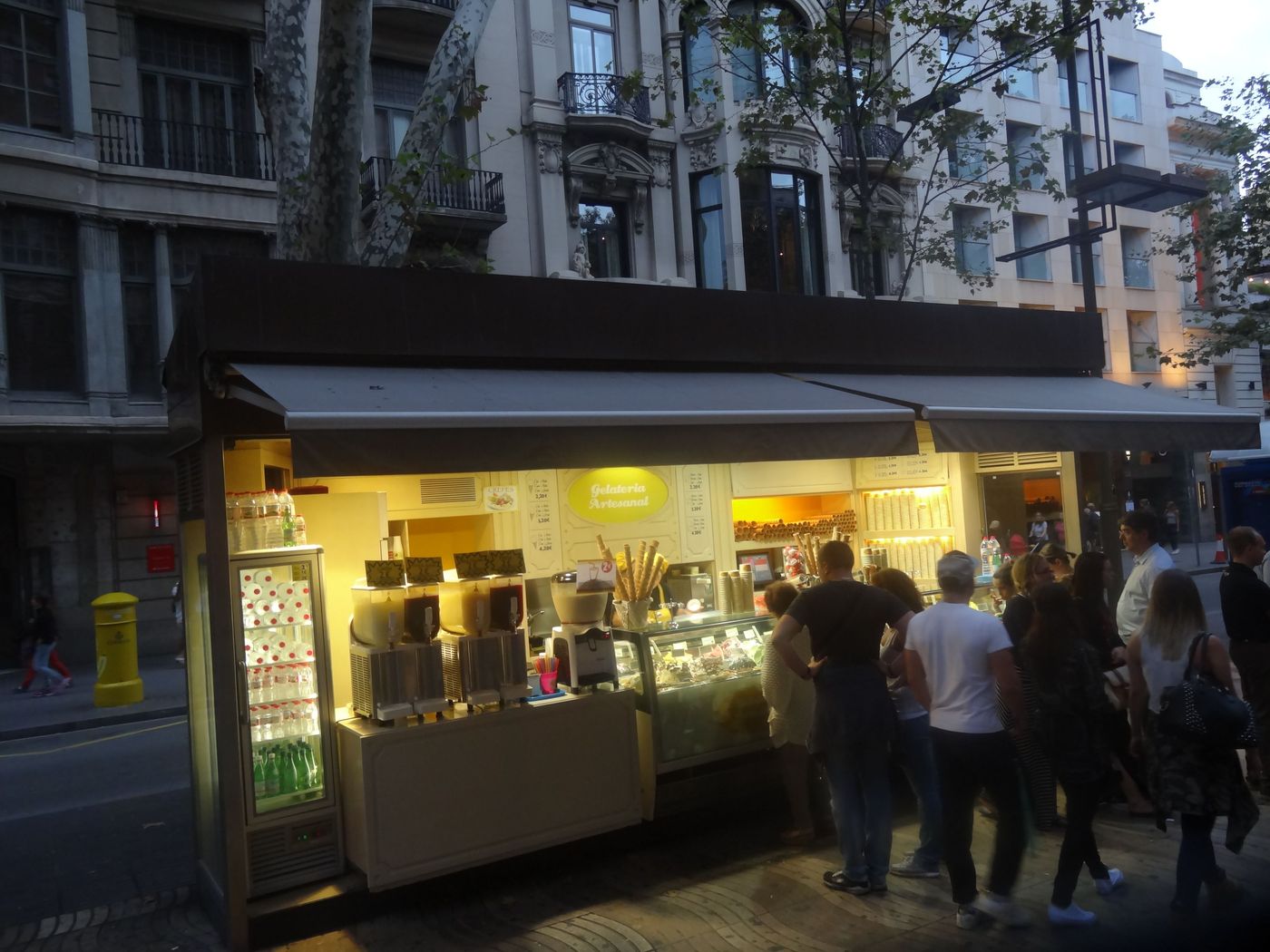 A lit ice-cream stand on La Rambla at dusk, people passing in front of it