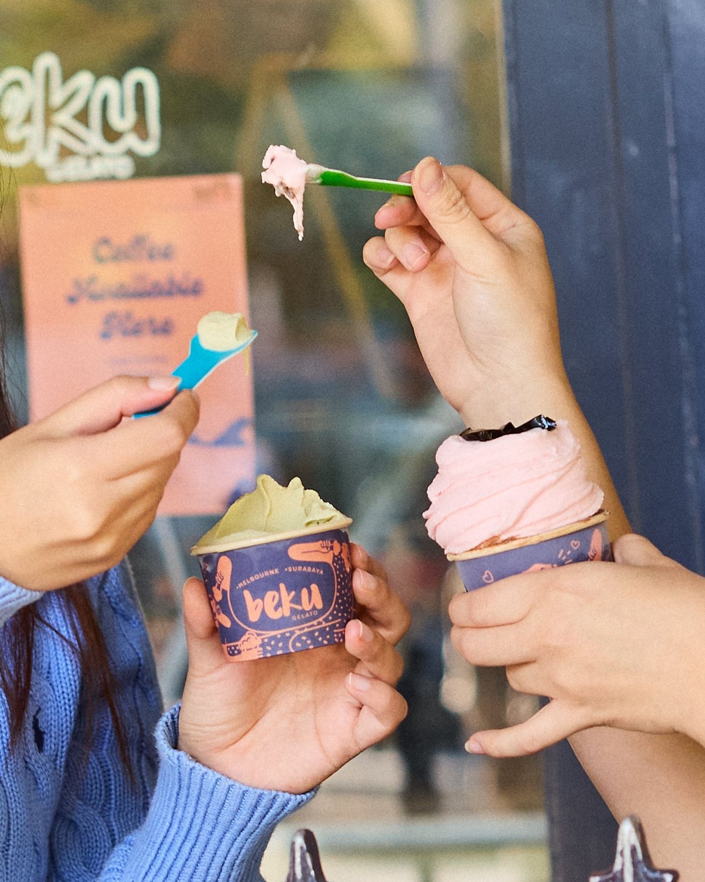 Two people outside the shop window holding purple Beku Gelato cups, one pale green and one pink, each lifting a coloured plastic spoon