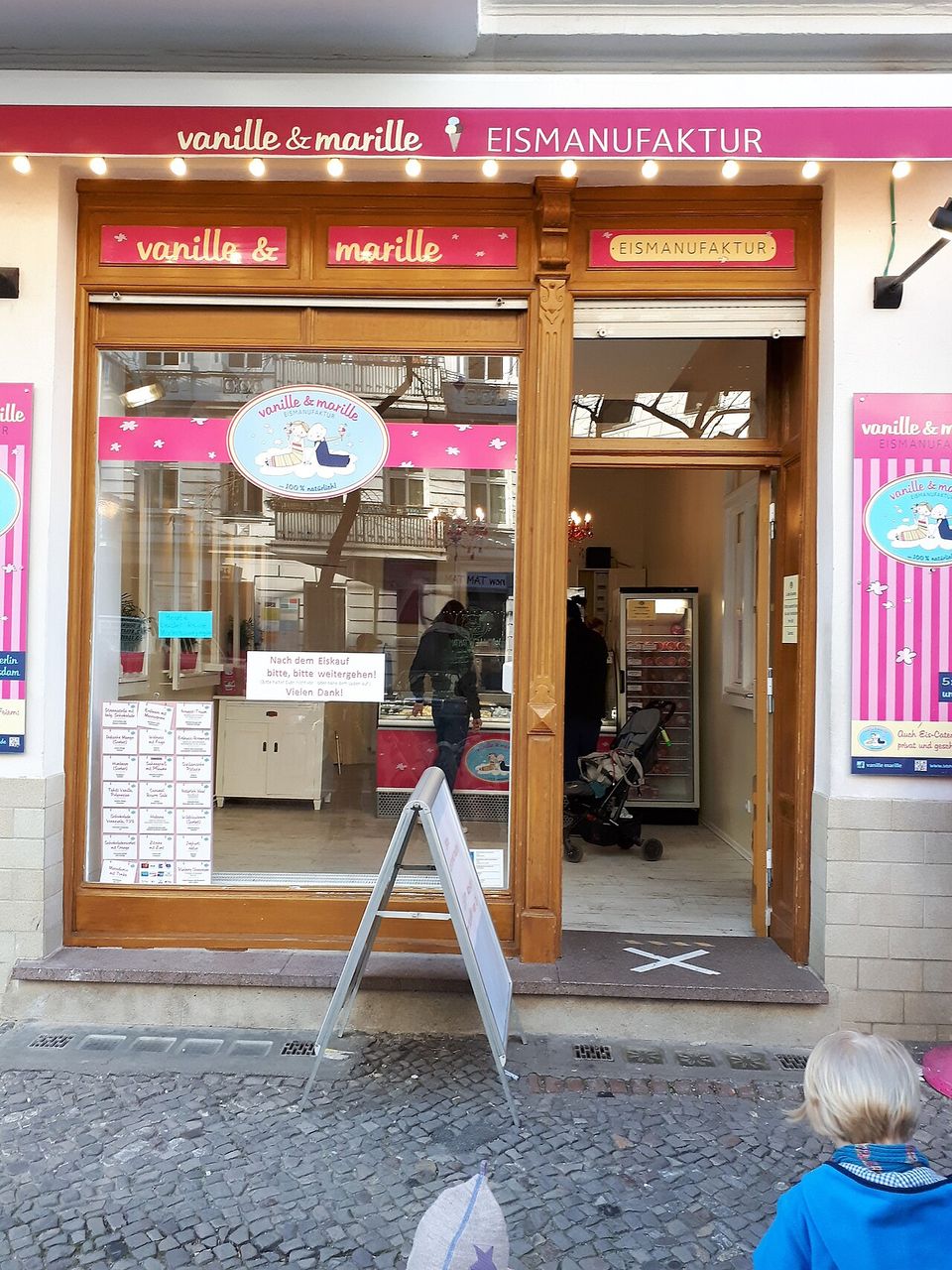 A pink and white shopfront lettered vanille & marille EISMANUFAKTUR, its flavour list taped up inside the window and a customer at the counter