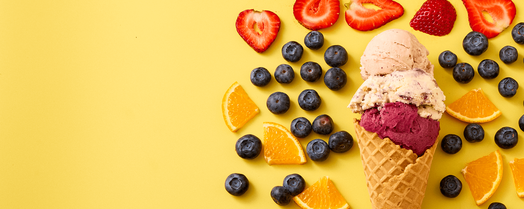 A cone and scoops of pale and berry-coloured ice cream among fresh strawberries, blueberries and orange segments