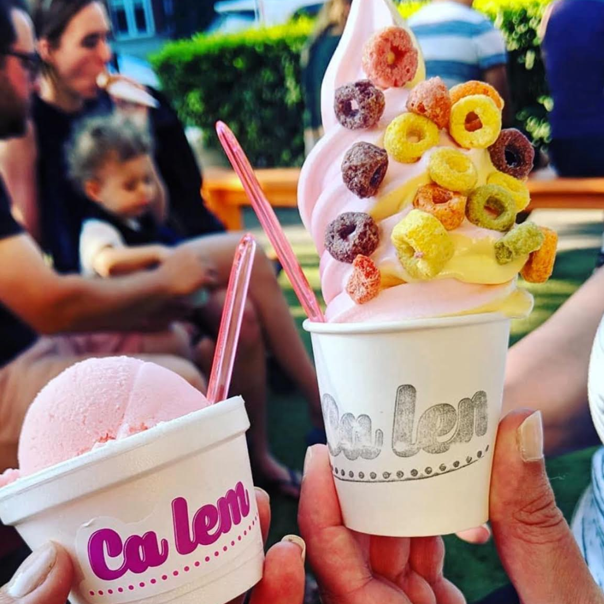 Hands holding cups of pink and cream soft serve piled with cereal and toppings