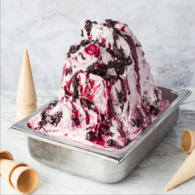 A metal tray heaped with pale pink gelato streaked with dark berry sauce and chocolate pieces, on marble with empty cones lying around it
