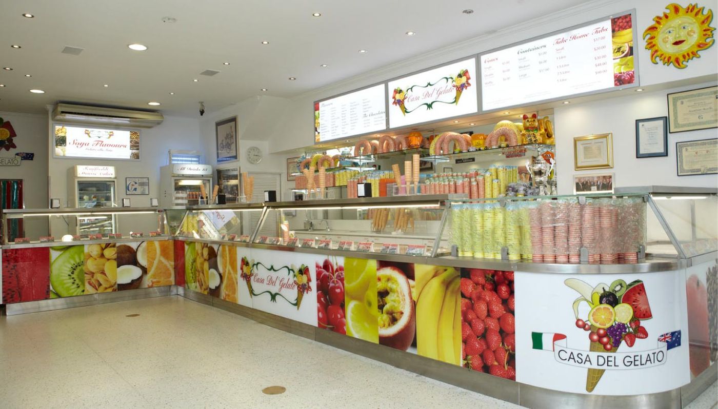 The L-shaped counter and display cases inside the shop, fruit panels along the front under the Casa del Gelato name, with lit menu boards and stacked cones behind