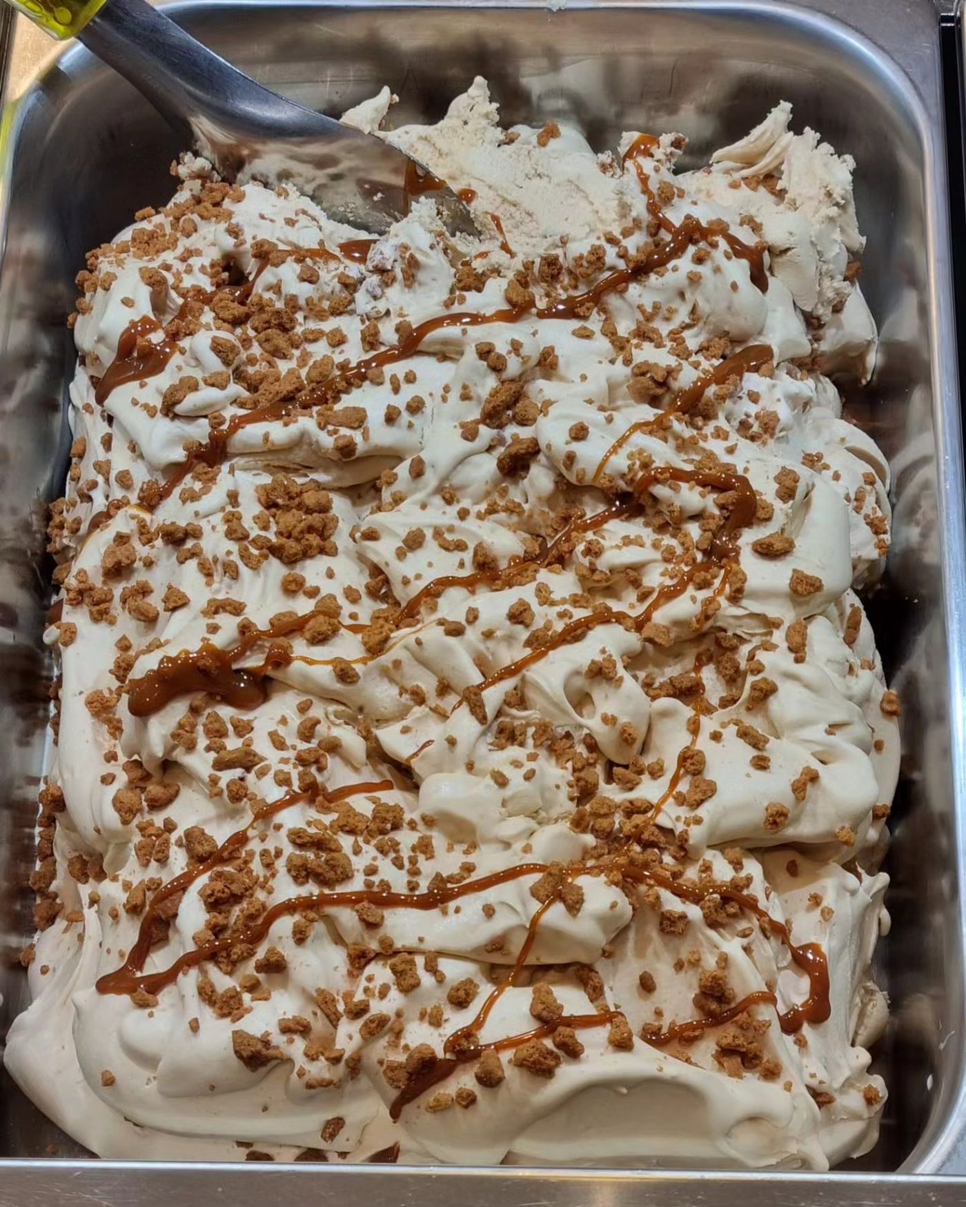 A steel pan of pale gelato drizzled with caramel and scattered with crumbled spiced biscuit, a spatula resting at one end
