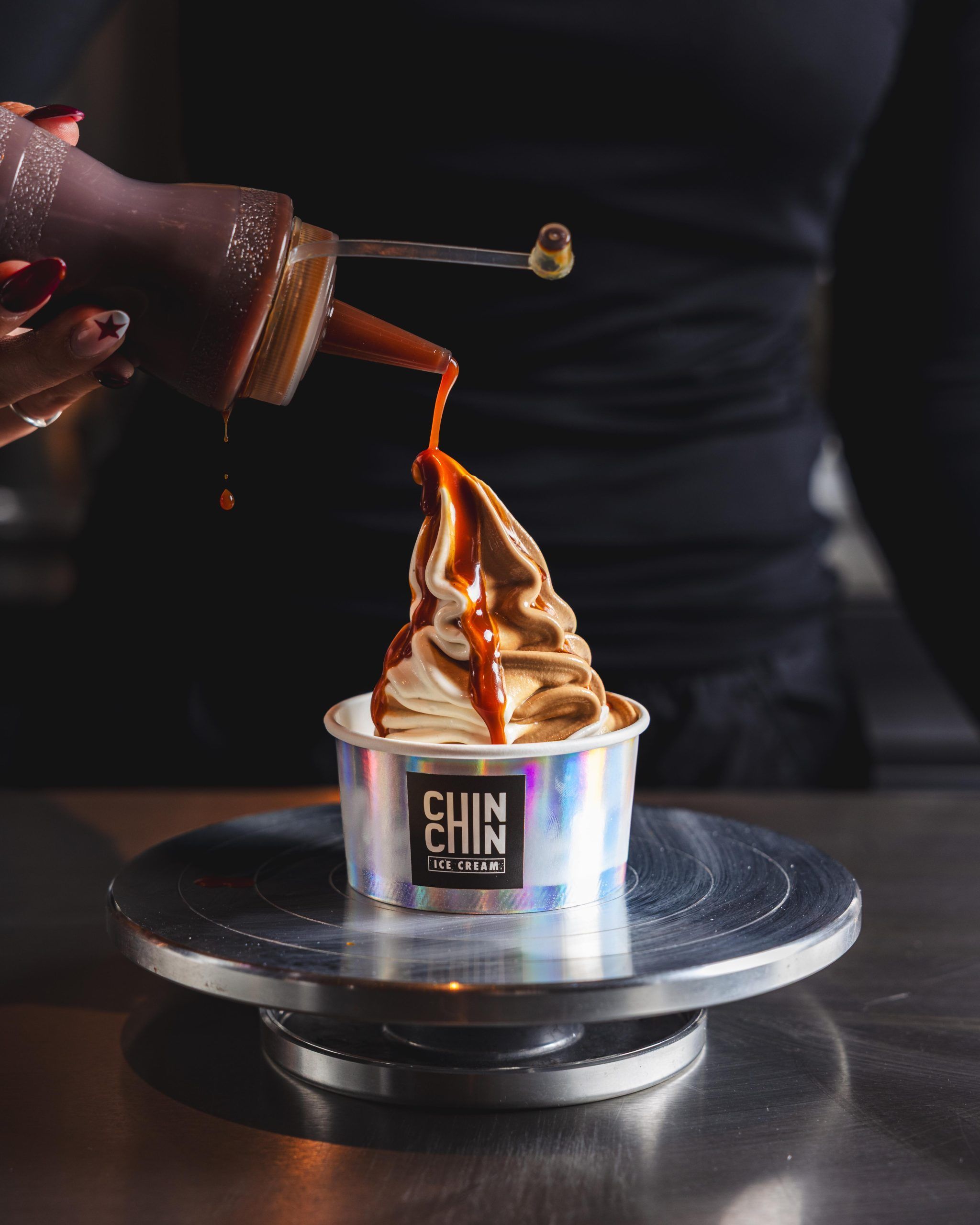 Chocolate sauce poured from a bottle over a swirl of soft serve in a branded cup, standing on a steel turntable