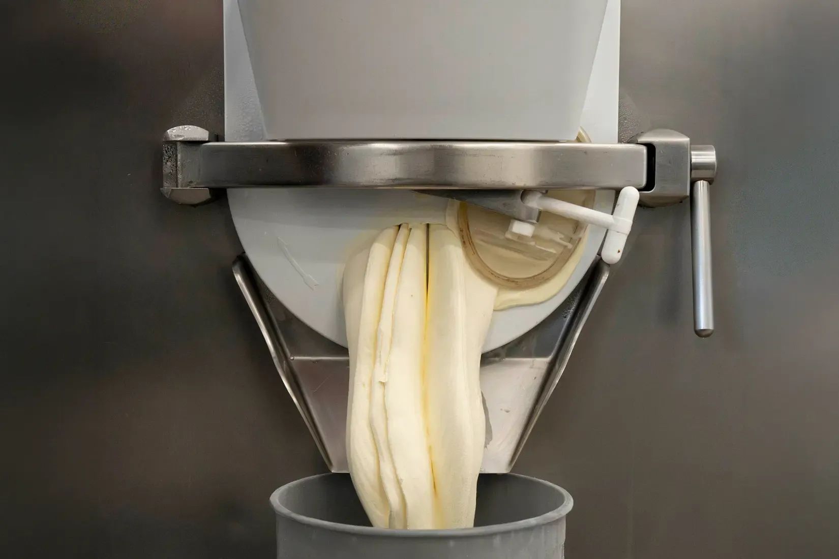 Pale gelato extruding in a thick ribbon from the spout of a batch freezer into a tub below