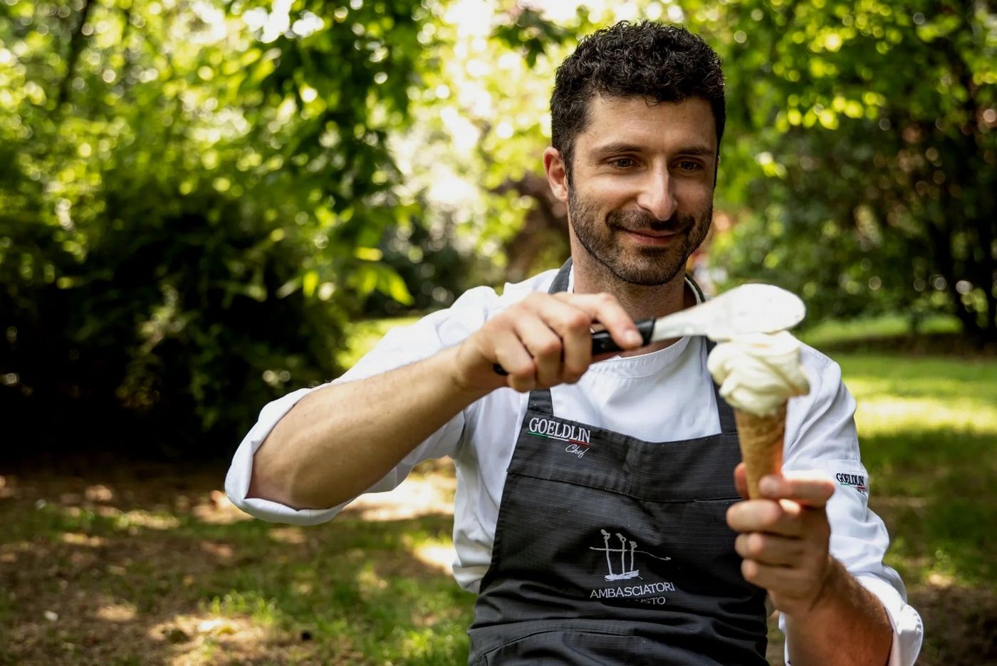 A man in chef's whites and a dark apron shaping pale gelato onto a cone with a spatula, outdoors among trees