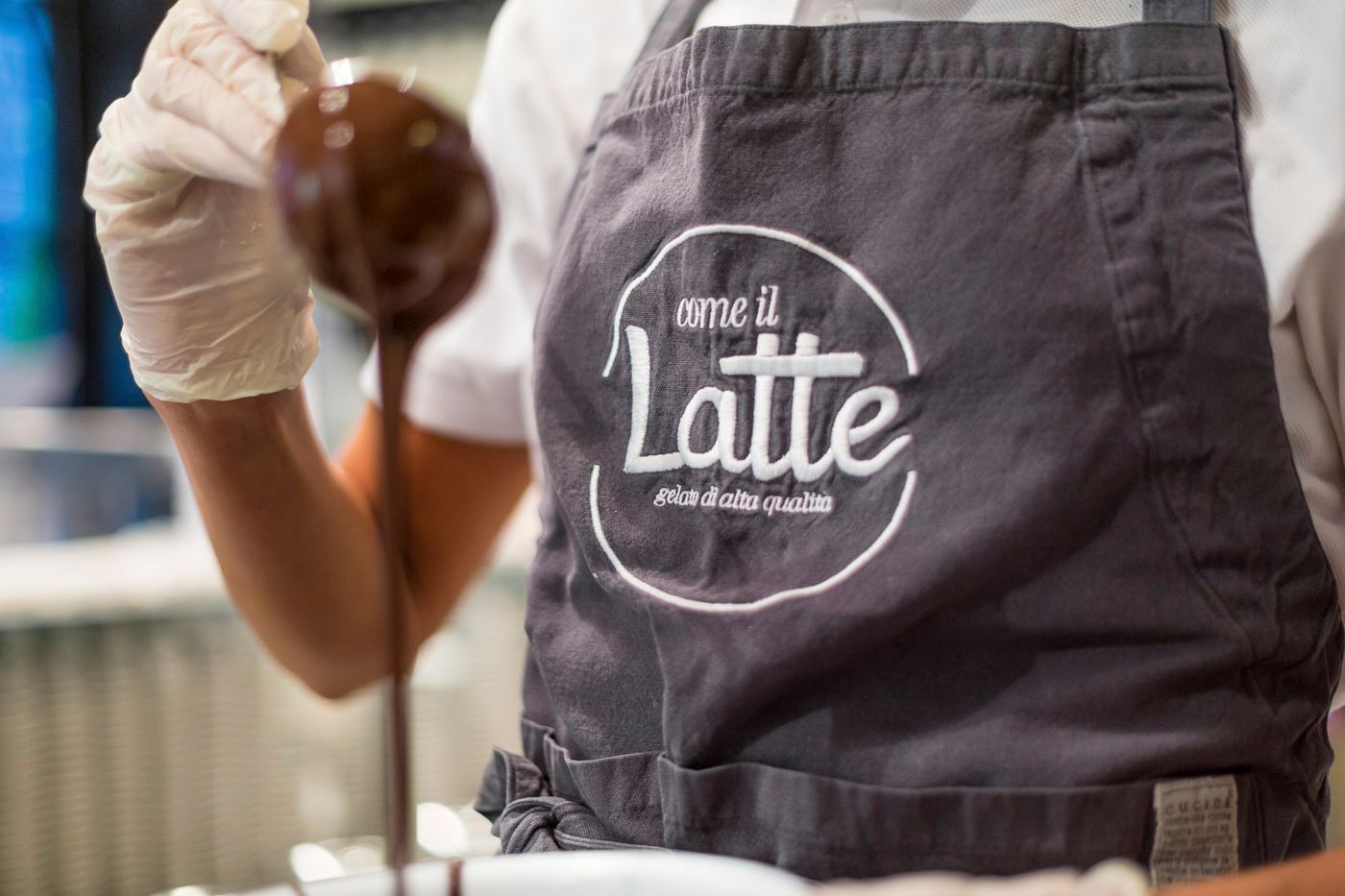 A gloved hand tipping dark chocolate from a ladle beside a grey apron embroidered with the Come il Latte roundel