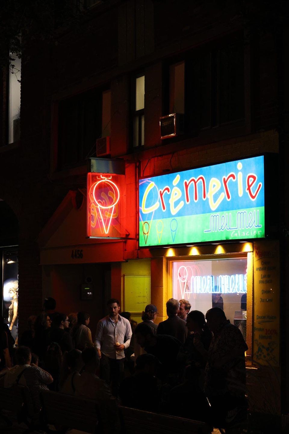 The lit Crémerie Meu Meu sign and a red neon cone above the service window at night, with people crowded on the pavement below