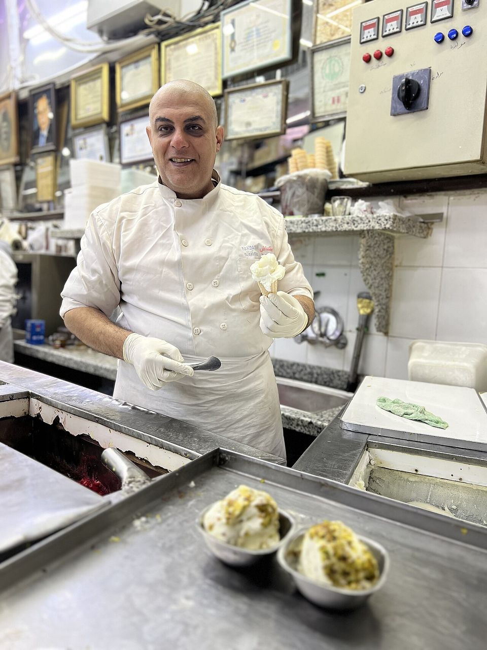 A man in chef's whites and gloves holding up a cone of pale booza behind steel trays, framed certificates crowding the wall above him