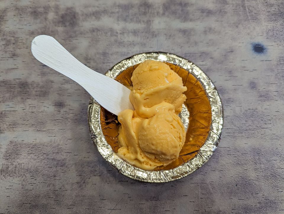 Two scoops of orange kulfi softening in a foil dish, a flat wooden spoon laid across it