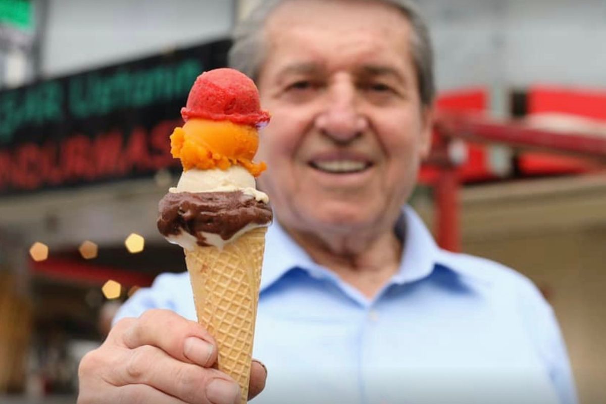 A man in a blue shirt holding out a tall cone of red and brown ice cream on a shopfront street