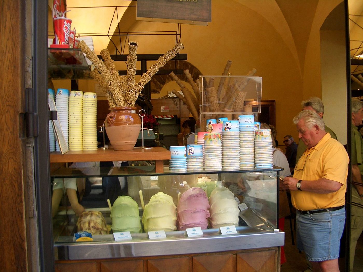 Mounds of pale gelato under a glass counter with cones stacked behind, a customer waiting to be served