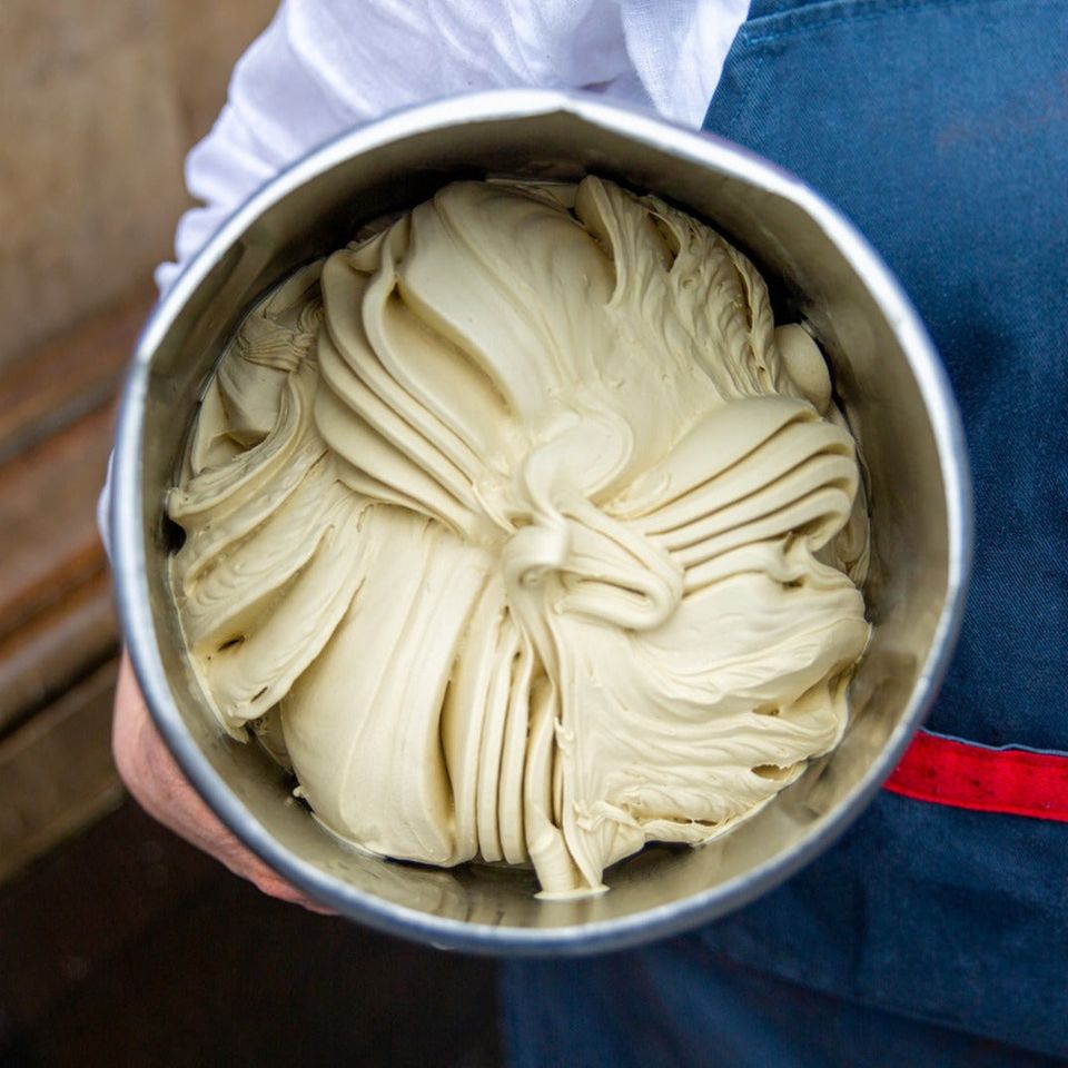 A stainless tub of pale pistachio gelato worked into ridges, carried against a blue apron and a white shirt sleeve