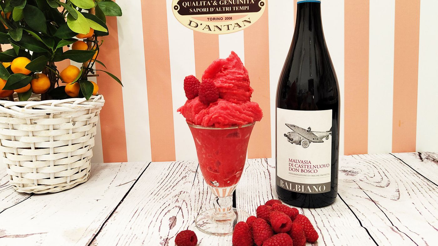A footed glass of raspberry sorbet heaped above the rim with whole raspberries on top, on a white plank table beside a bottle of Malvasia di Castelnuovo Don Bosco and a potted citrus tree