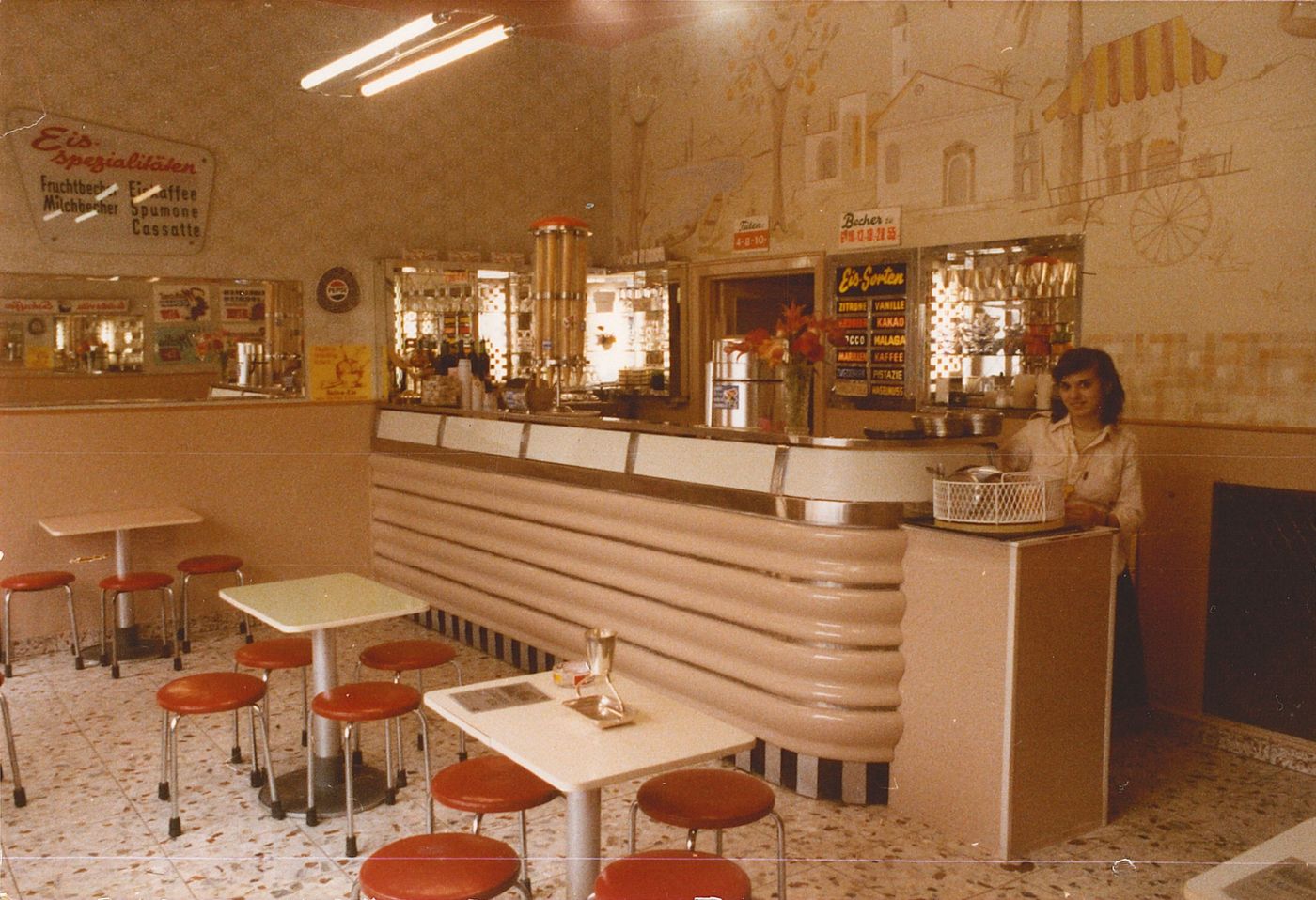 An old colour photograph of the shop interior, with a ribbed white counter, red vinyl stools at formica tables, a painted Italian street scene on the wall and a flavour board listing Zitrone, Vanille and Pistazie