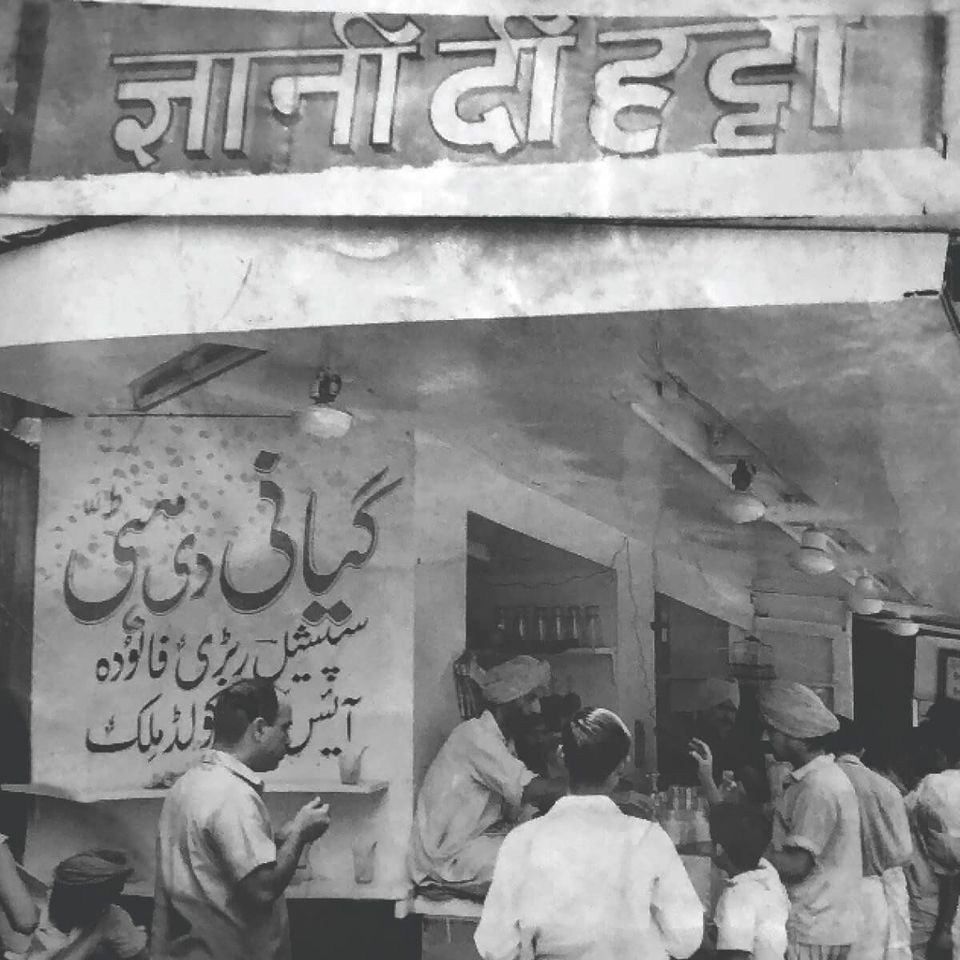 A black and white archive photograph of the shopfront, its Urdu signboard above men working behind an open counter