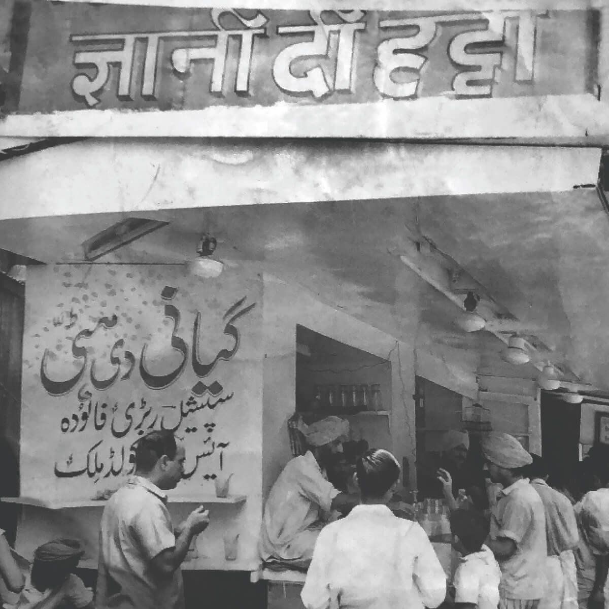 A black and white archive photograph of the shopfront, its Urdu signboard above men working behind an open counter