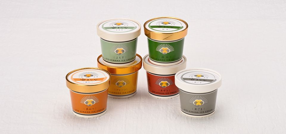Six lidded tubs of Kyo Kinana ice cream arranged in two rows on white cloth, their lids banded in green, gold, orange, red and grey