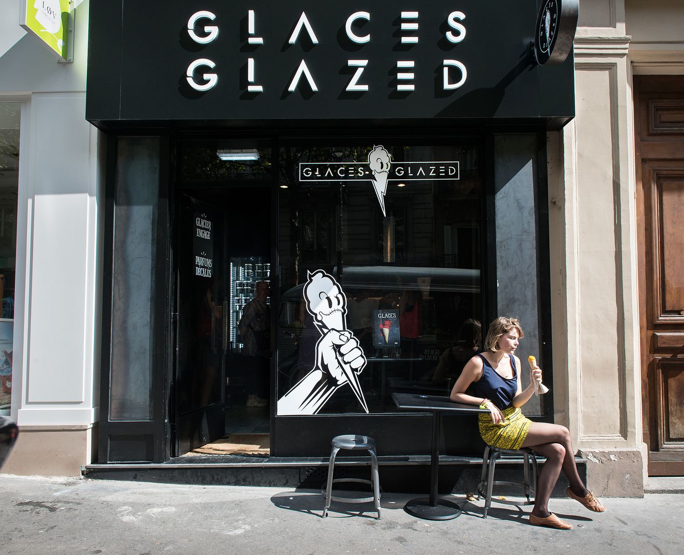 The black shopfront at 54 Rue des Martyrs under GLACES GLAZED lettering, a cartoon fist holding a cone painted on the window, and a woman on a stool at the pavement table eating an ice cream