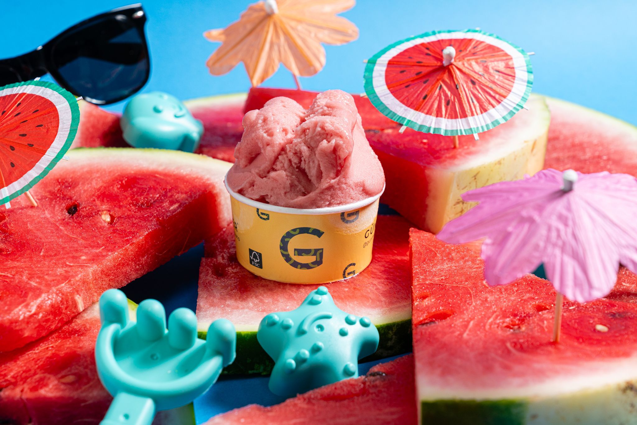 A scoop of pink gelato in a yellow cup, set on watermelon slices among paper cocktail parasols