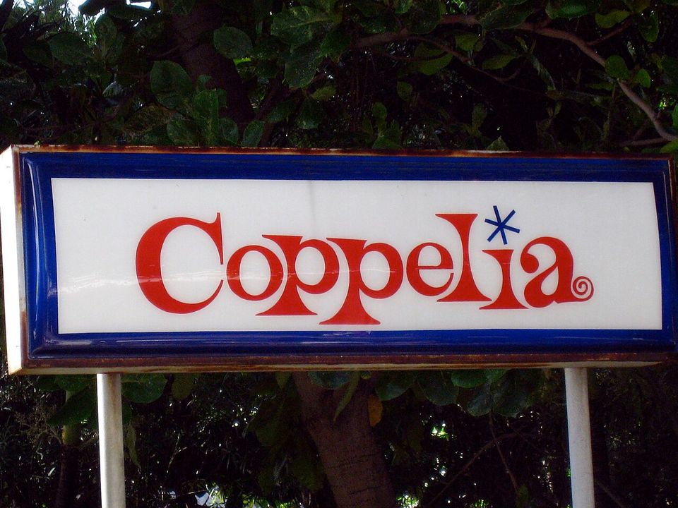The Coppelia sign: red script lettering with a blue star above the i, on a white panel in a rusting blue frame, mounted on posts against dark foliage