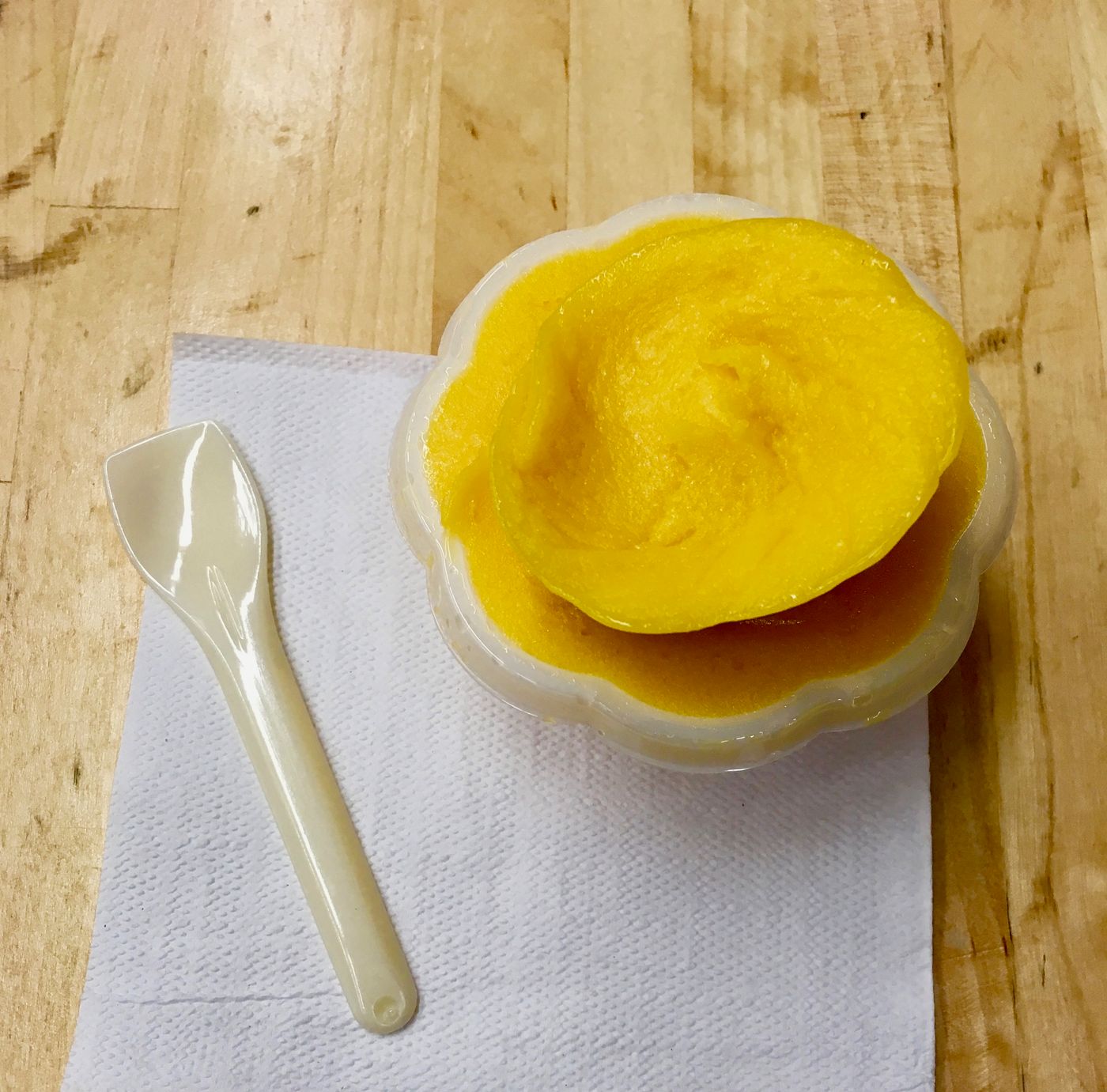 A scoop of bright yellow sorbet in a fluted plastic cup on a paper napkin, a white plastic spoon laid beside it on a wooden counter