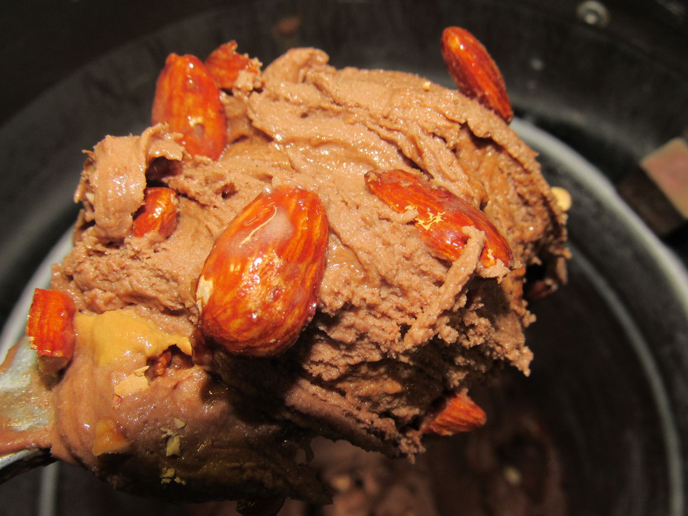 A spade lifting chocolate ice cream studded with whole glazed almonds out of a metal tub