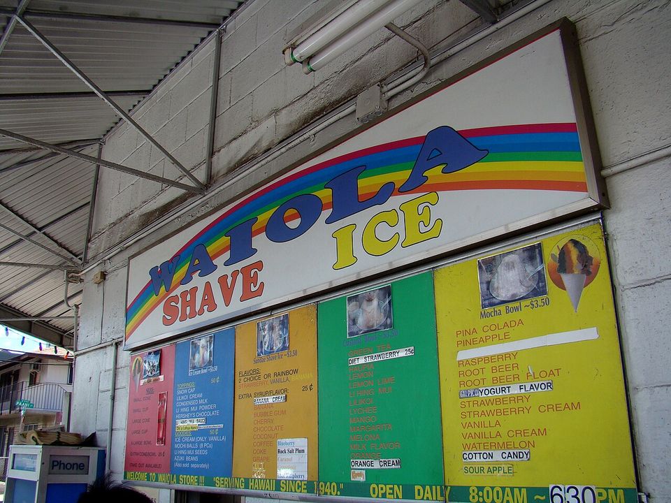 A rainbow-lettered WAIOLA SHAVE ICE sign above five colour-coded boards of flavours and prices, and a strip reading SERVING HAWAII SINCE 1940