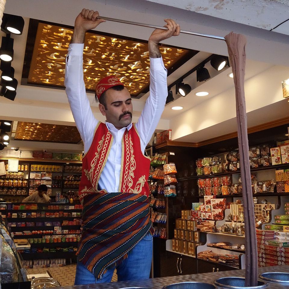 A vendor in a red embroidered waistcoat and cap stretching a long rope of ice cream down from a rod held above his head