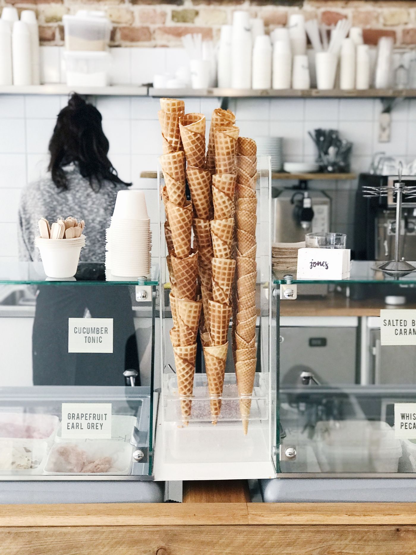 Waffle cones stand upright in a clear acrylic holder at the centre of a glass ice-cream counter, with tubs labelled Cucumber Tonic and Grapefruit Earl Grey to the left and shelves of white cups on the brick wall behind