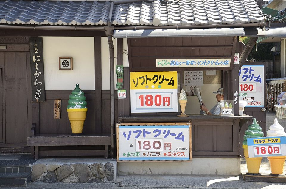 A soft-serve stall built into a wooden shopfront, hand-lettered 180円 signs on every side and green matcha cone models standing about, the vendor at the hatch in a cap