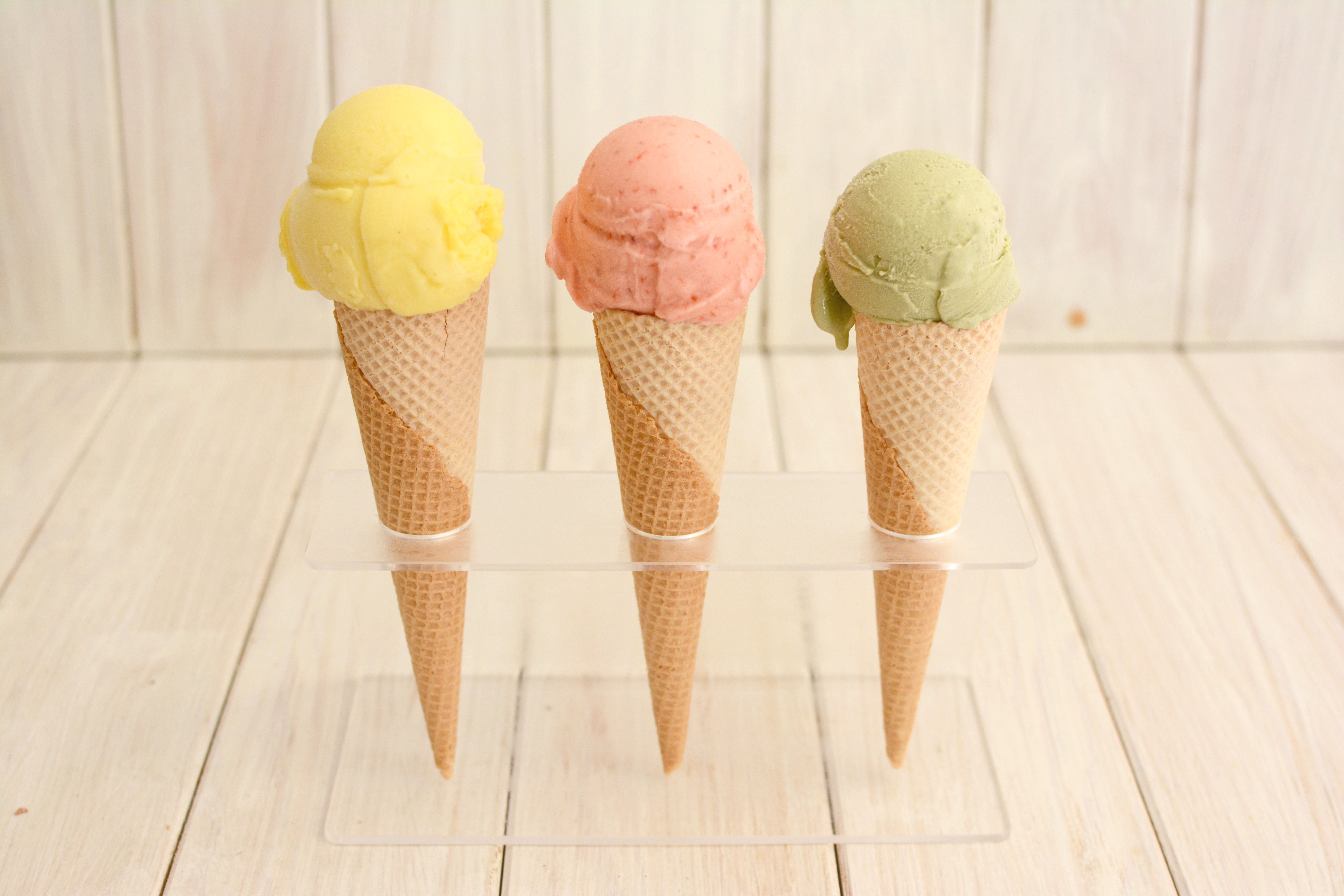 Three ice cream cones with yellow, pink and green scoops standing in a stand against a pale wooden backdrop