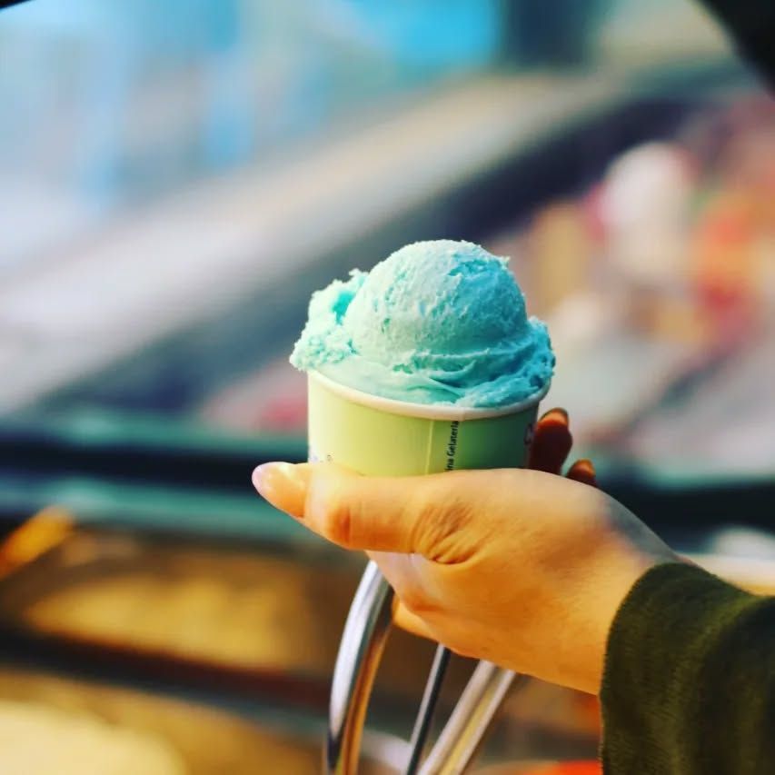 A hand holding a small tub of pale blue ice cream over a display counter