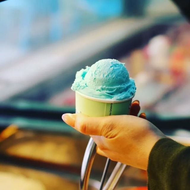 A hand holding a small tub of pale blue ice cream over a display counter
