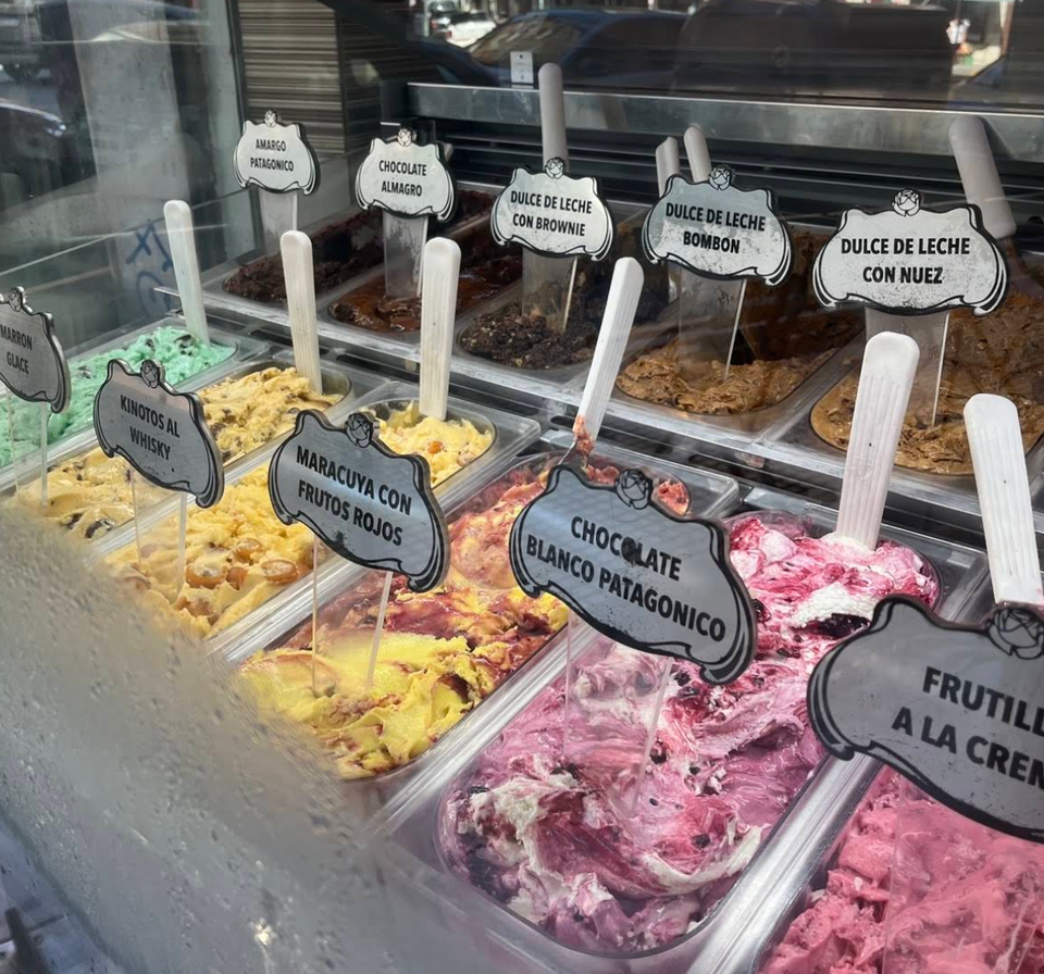Pans of ice cream behind the display-case glass with a white spade standing in each, labelled Chocolate Almagro, Dulce de Leche con Brownie, Dulce de Leche Bombón, Dulce de Leche con Nuez, Maracuyá con Frutos Rojos, Chocolate Blanco Patagónico and Kinotos al Whisky