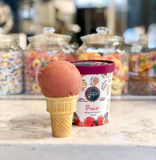 A cone holding one pink scoop stands on a marble counter beside a 500ml tub labelled Fraise in Bilboquet livery, with blurred glass jars of coloured sweets on the shelf behind