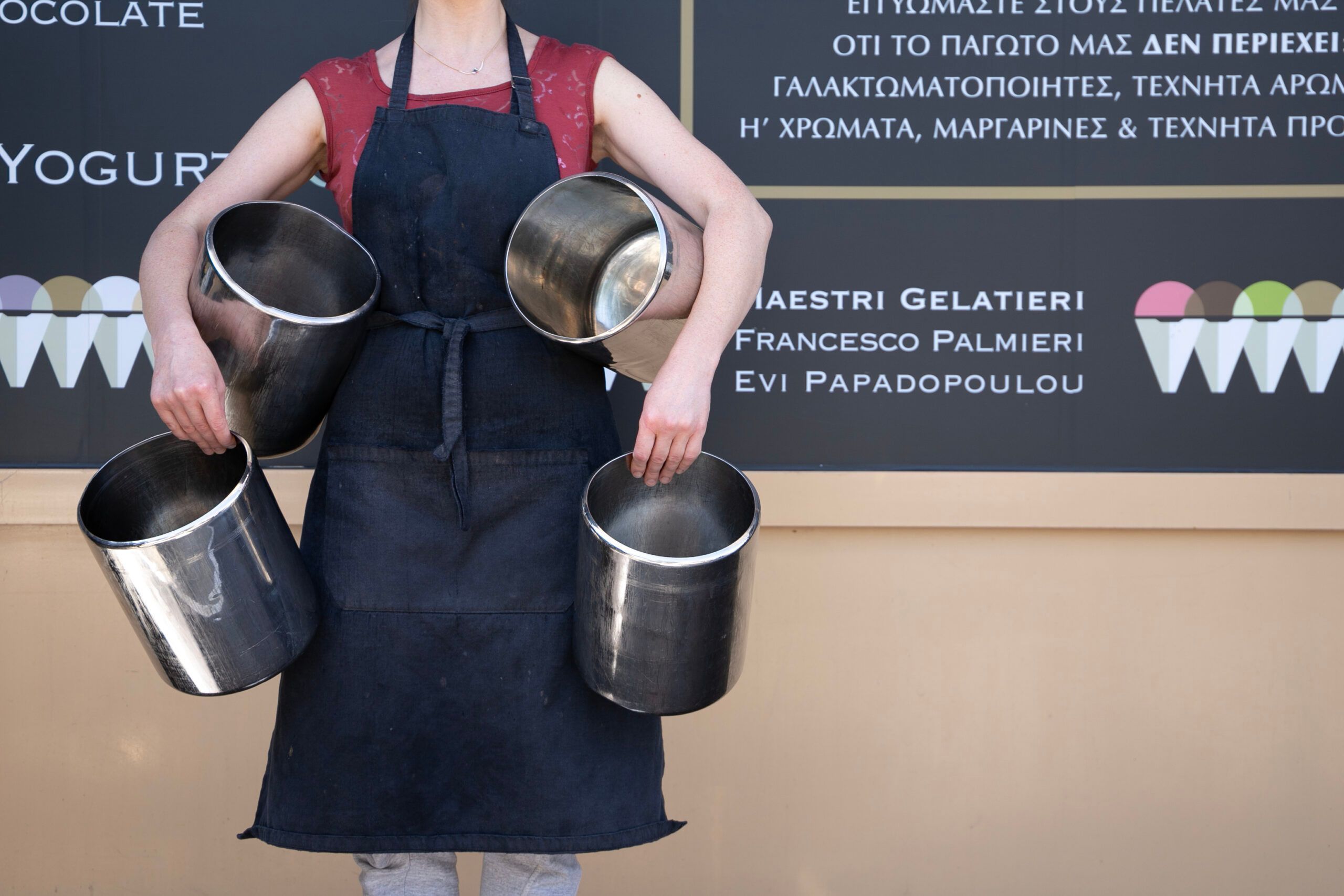 A person in a dark apron carrying four steel gelato canisters, two in each hand