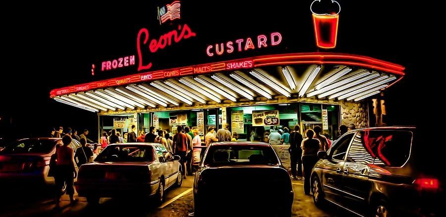The neon-lit drive-in at night, cars pulled up under the Leon's Frozen Custard sign