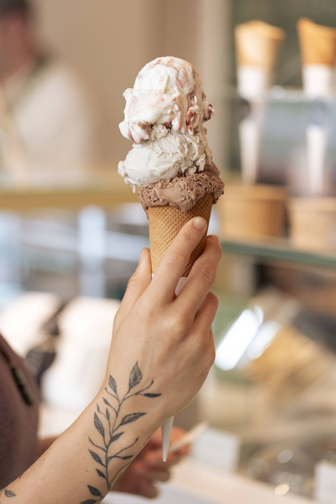 A hand with a leafy-branch tattoo on the forearm holds a waffle cone carrying three scoops — chocolate at the bottom, pale cream in the middle, white streaked with red on top — in front of a blurred counter