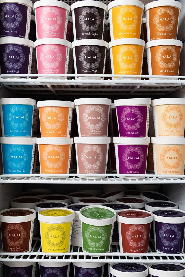 Pints in coloured Malai livery stand in rows on the wire shelves of an open freezer, labelled Sweet Milk, Masala Chai, Turkish Coffee, Sea Salt Vanilla, Orange Fennel, Star Anise, Carrot Halwa, Lemon Cardamom, Toasted Nutmeg and Ginger Root
