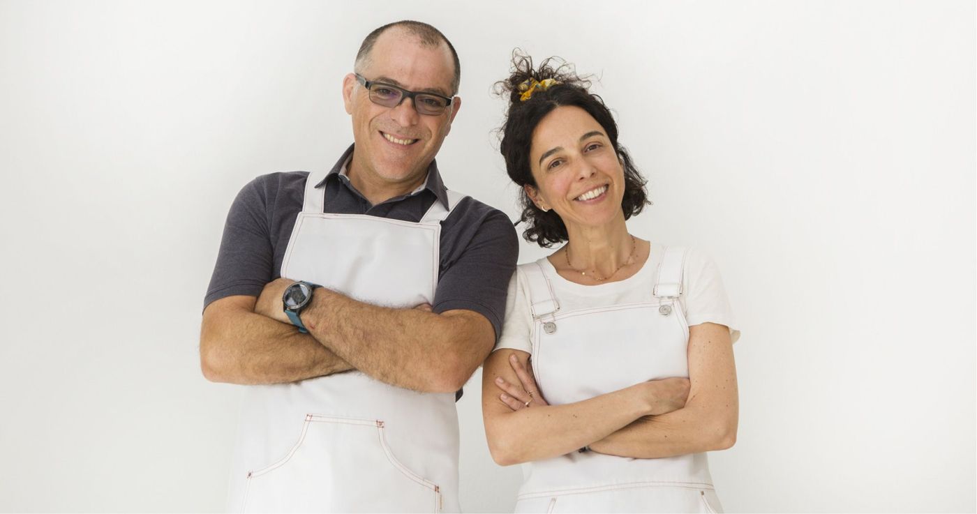 Two ice-cream makers in white aprons standing side by side against a plain pale wall, arms folded